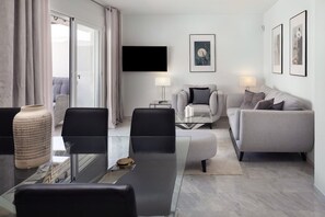 A modern living room with a glass dining table, grey sofas, a flat-screen TV, and artwork on the wall.