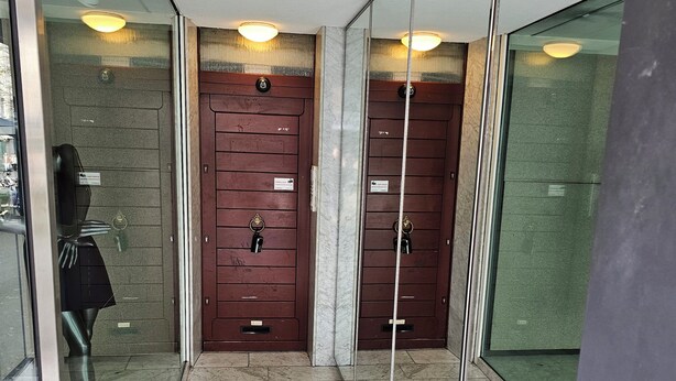 Un pasillo con dos puertas de madera, cada una con cerradura y un cartel arriba. El suelo está cubierto de baldosas y hay una pared de vidrio a la izquierda.
