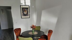 Un comedor con una mesa redonda, cuatro sillas, un jarrón con flores, un cuadro enmarcado en la pared y una lámpara colgante de diseño geométrico.