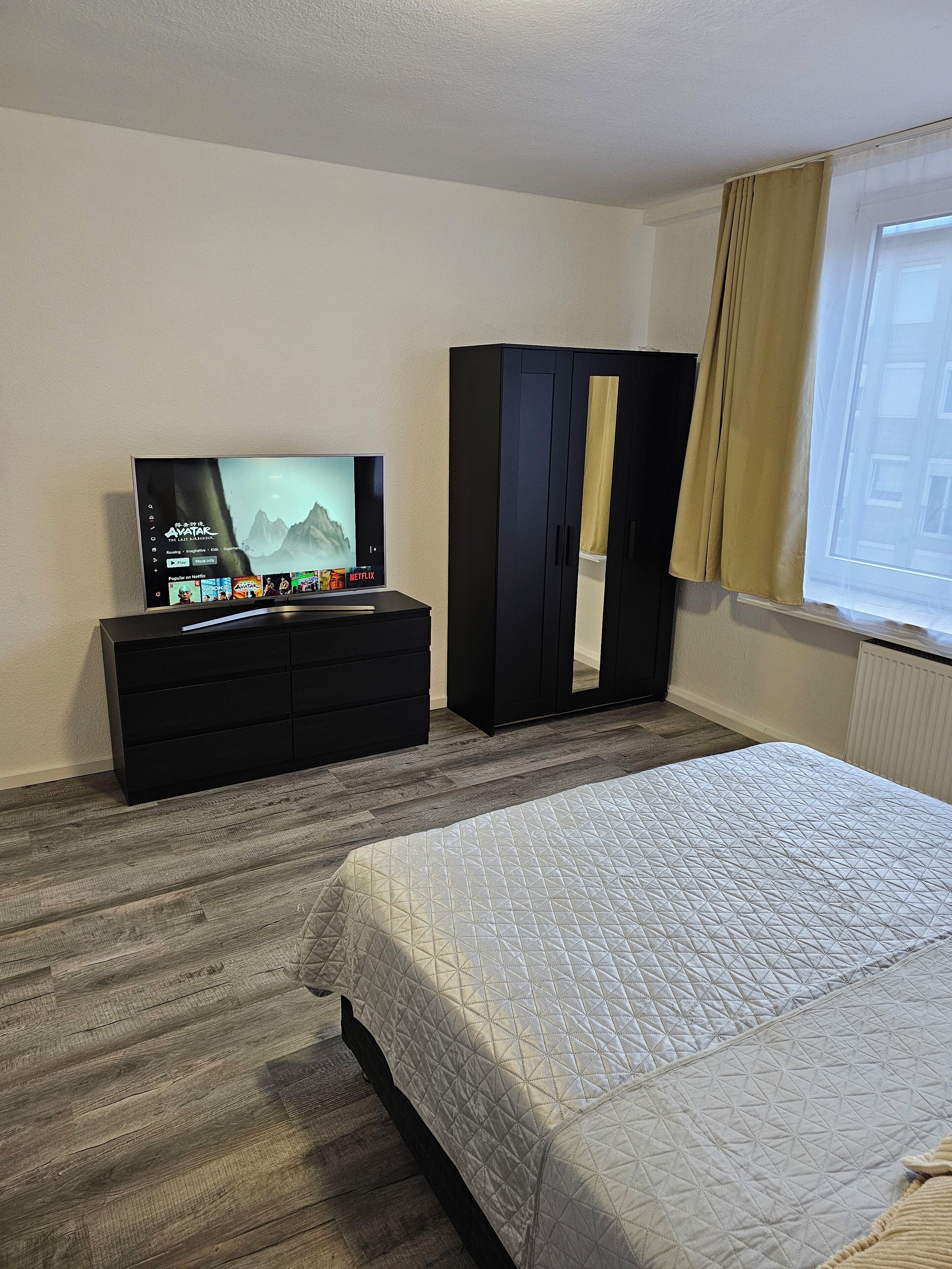 Una habitación de hotel con una cama, un televisor en una cómoda, un armario y una ventana con cortinas.