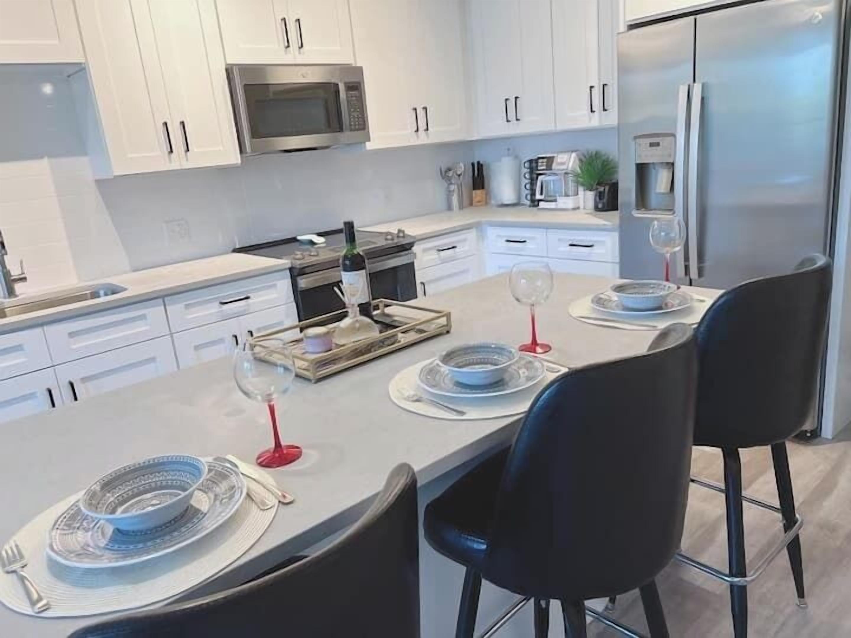 A modern kitchen with white cabinets, stainless steel appliances, and a dining area set for two with black chairs and a tray of dishes.
