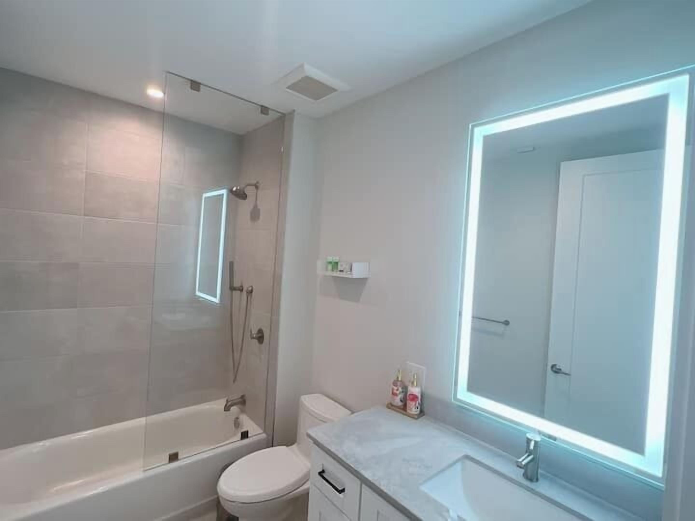 A modern bathroom with a bathtub, toilet, and a large mirror with built-in lighting.