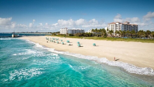A beach with clear turquoise water, a sandy shore, and several umbrellas. There are buildings in the background. - Singer Island | Coastal Escape | Pools & Beach Days (Palm Beach Shores)