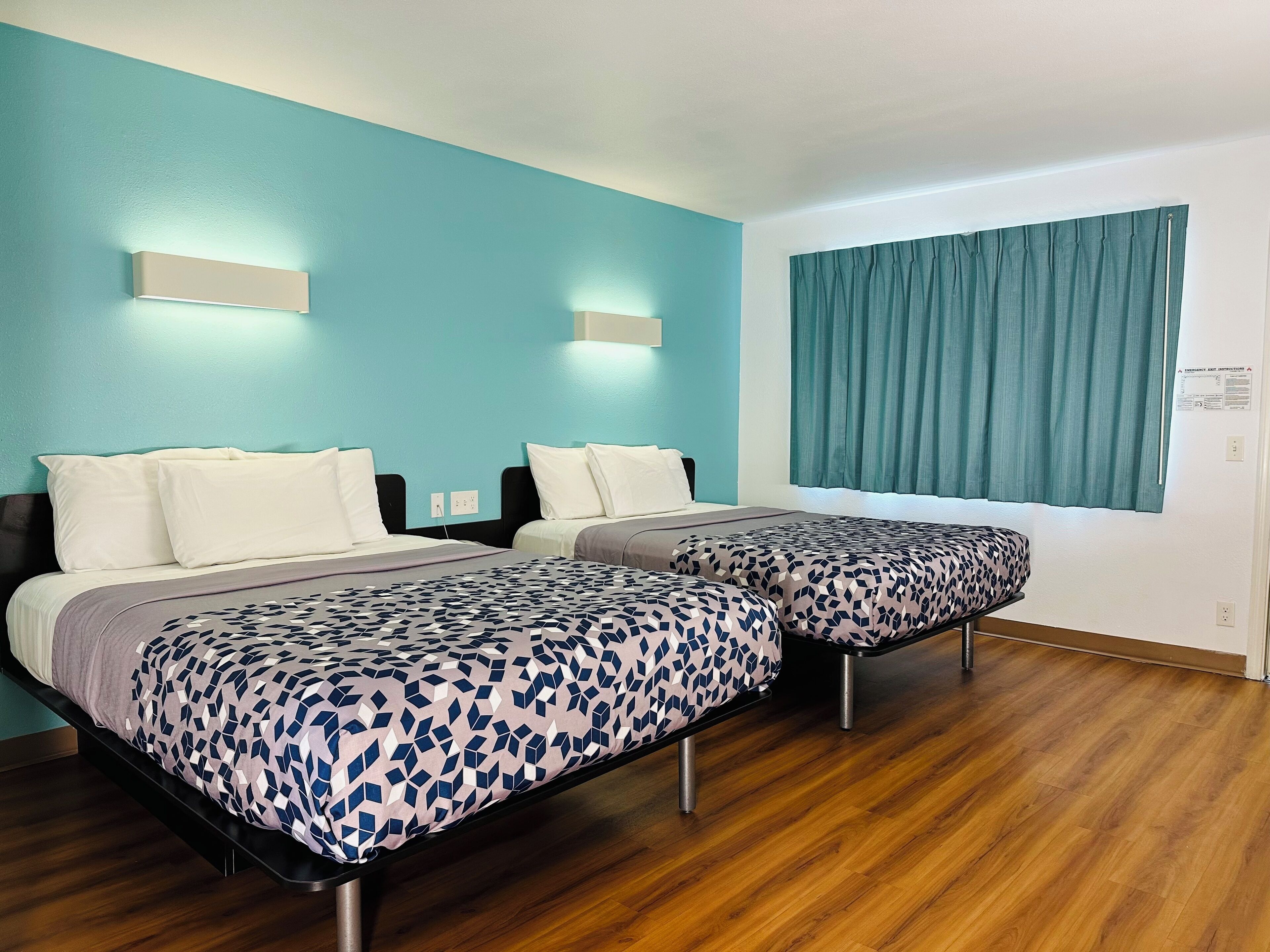 A hotel room with two beds, teal walls, wooden flooring, and a window with curtains.