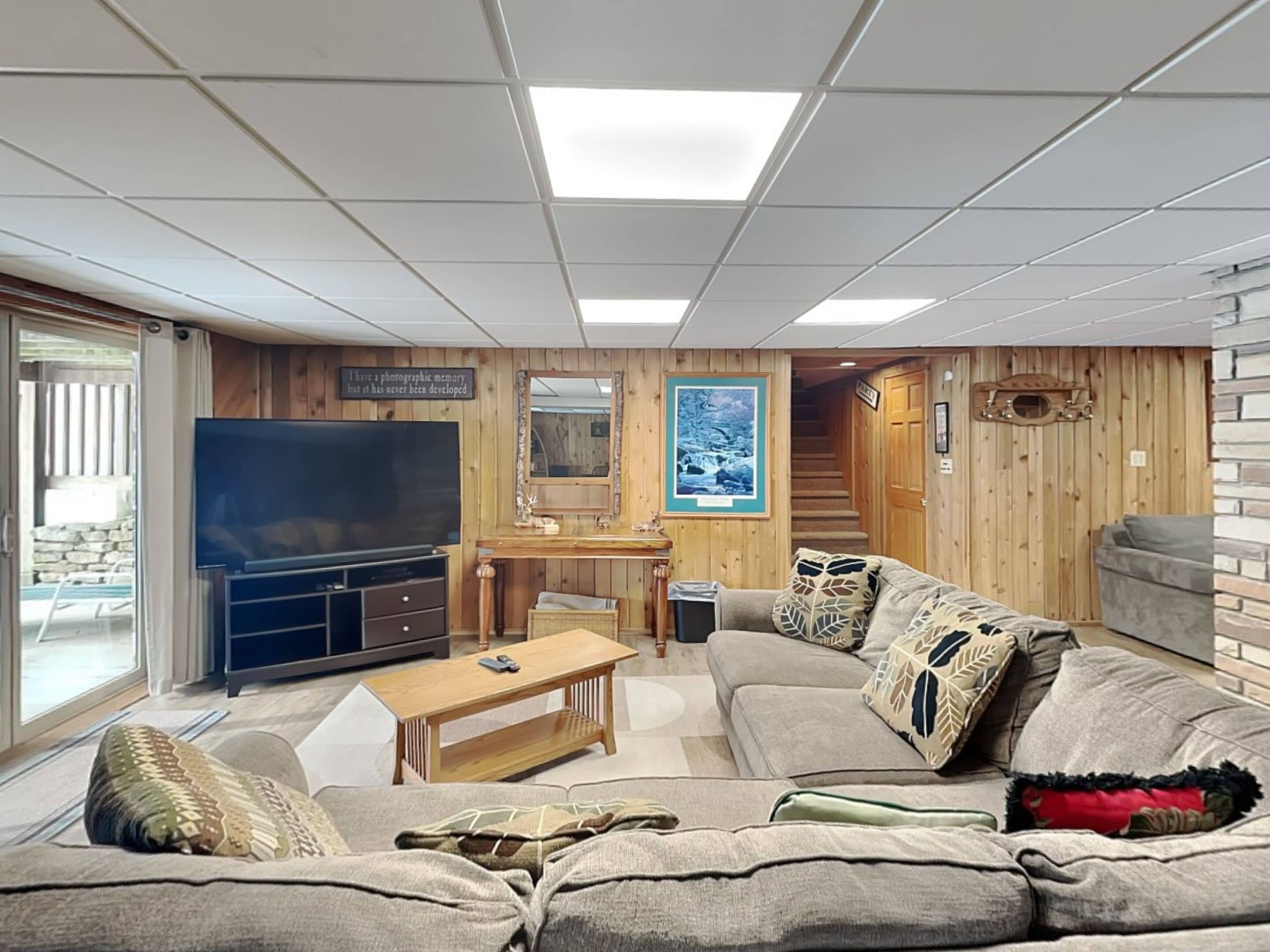 A cozy living room with a large flat-screen TV, wooden panel walls, a staircase, and a comfortable sofa.