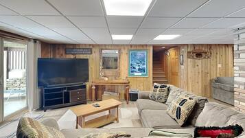 A cozy living room with a large flat-screen TV, wooden panel walls, a staircase, and a comfortable sofa.