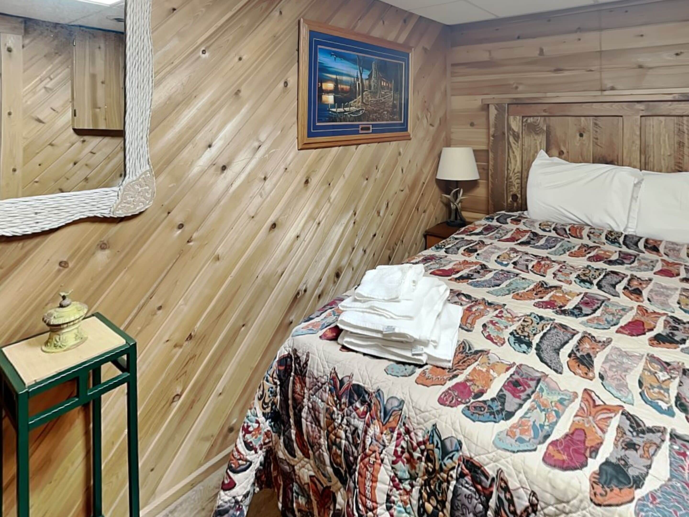 A wooden-paneled room with a bed, a framed picture on the wall, a lamp, and a small table with a decorative item.