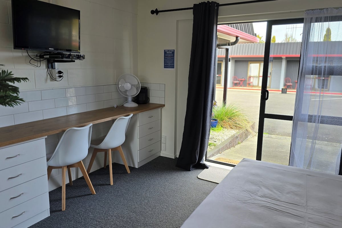 A hotel room with a bed, a desk with two chairs, a TV, a fan, and a view of the outside.