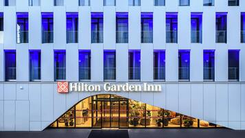 Welcome to the Hilton Garden Inn, featuring a modern design with a grid-patterned facade and a warm entrance area with potted plants and outdoor seating.