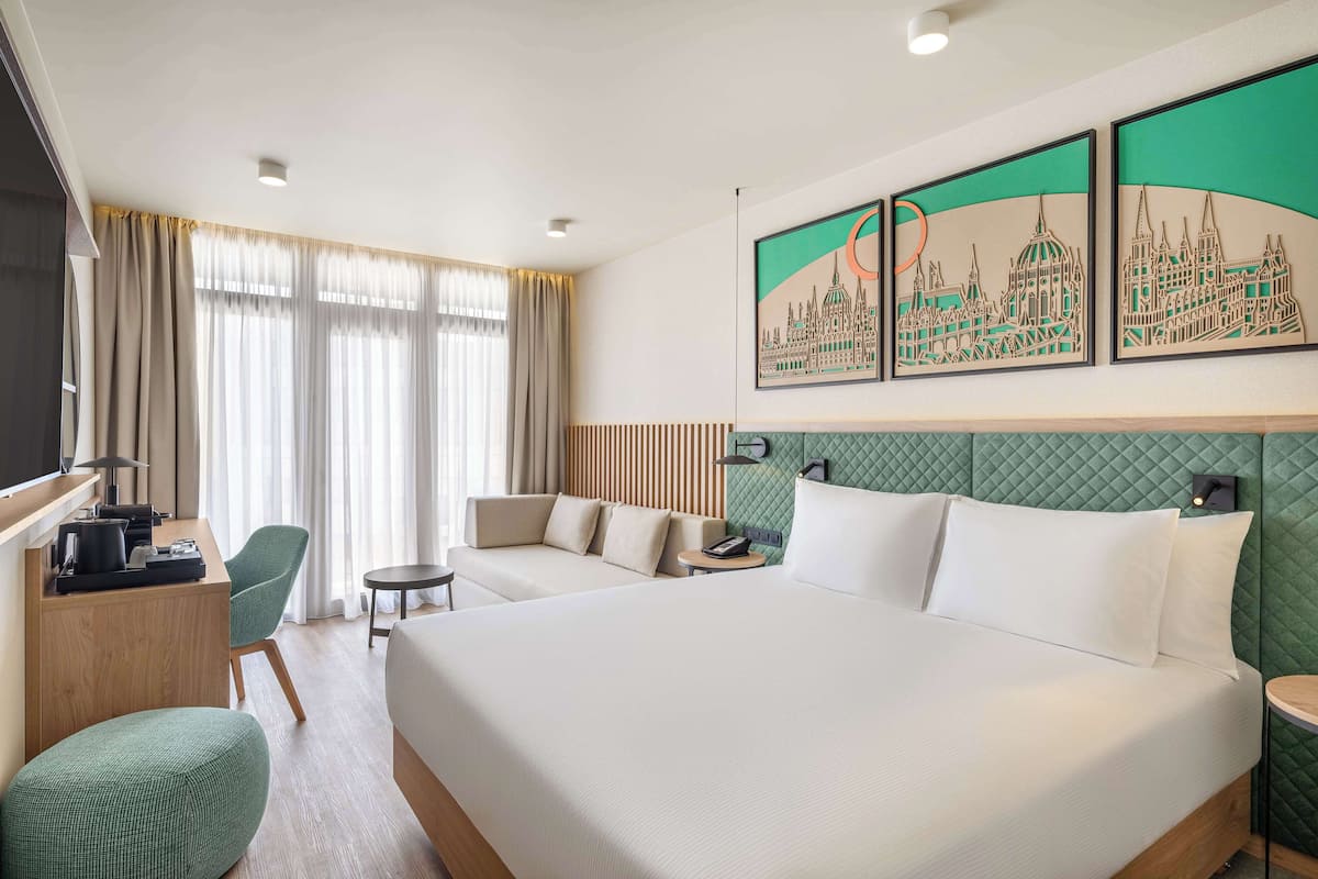 Habitación de hotel con una cama grande, dos zonas de estar, un escritorio con computadora y obras de arte que representan un paisaje urbano.