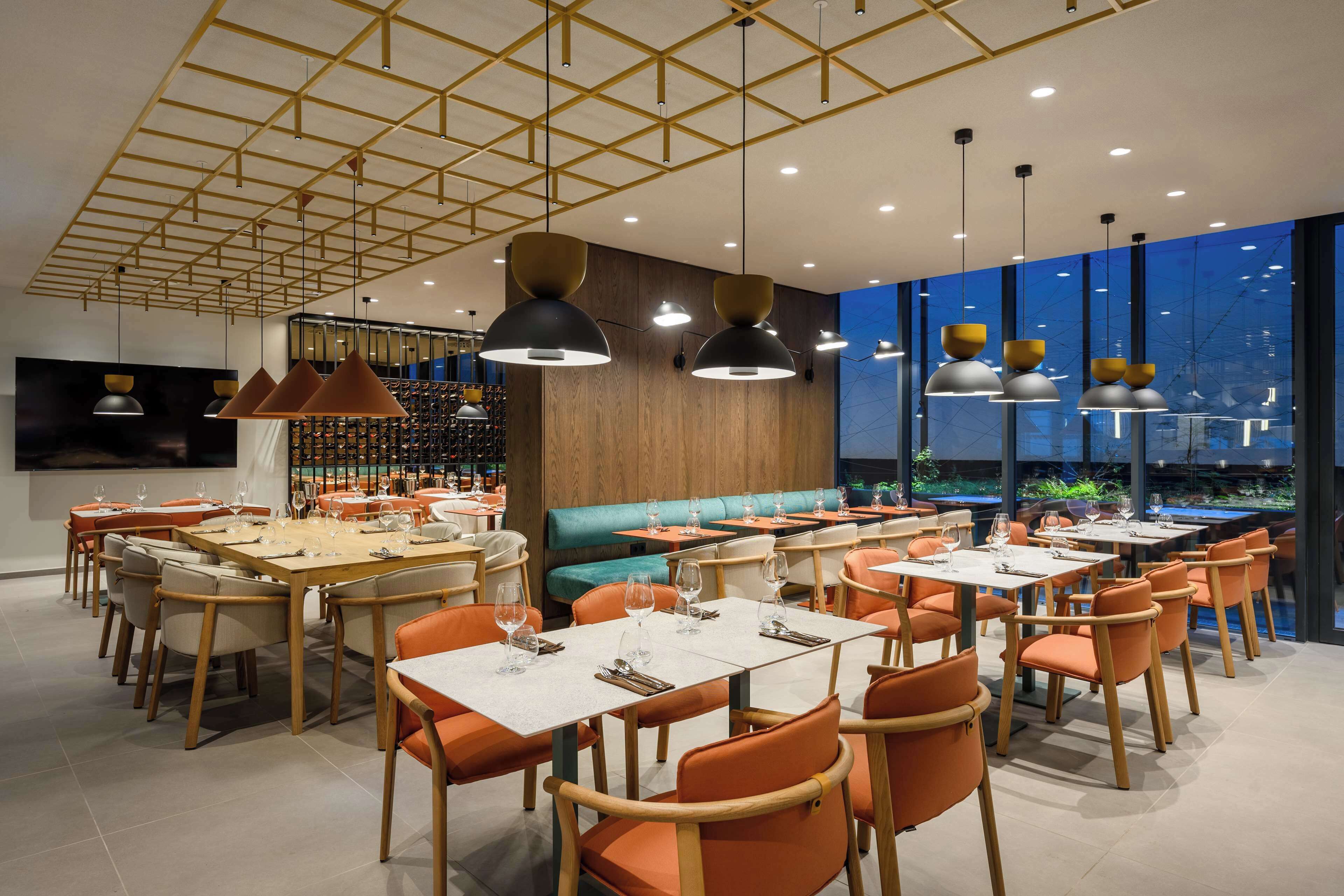 A modern restaurant with multiple dining tables and chairs, a wooden accent wall, and pendant lighting.