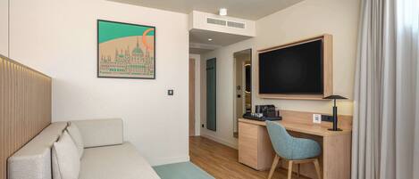 A hotel room with a sofa, a desk, a chair, a flat-screen TV, and a painting on the wall.