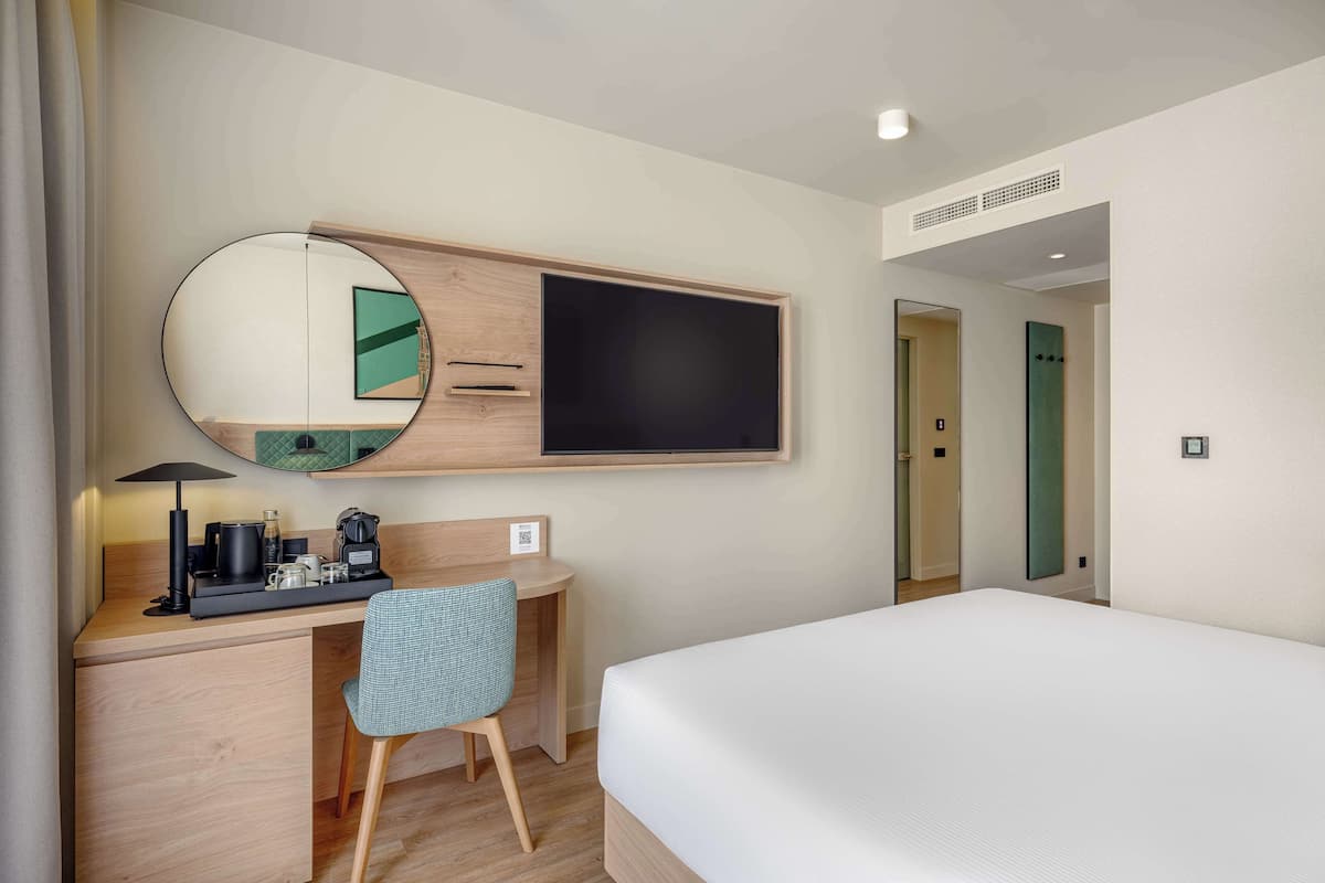Una habitación de hotel con una cama, un escritorio con una silla, un espejo redondo y un televisor.