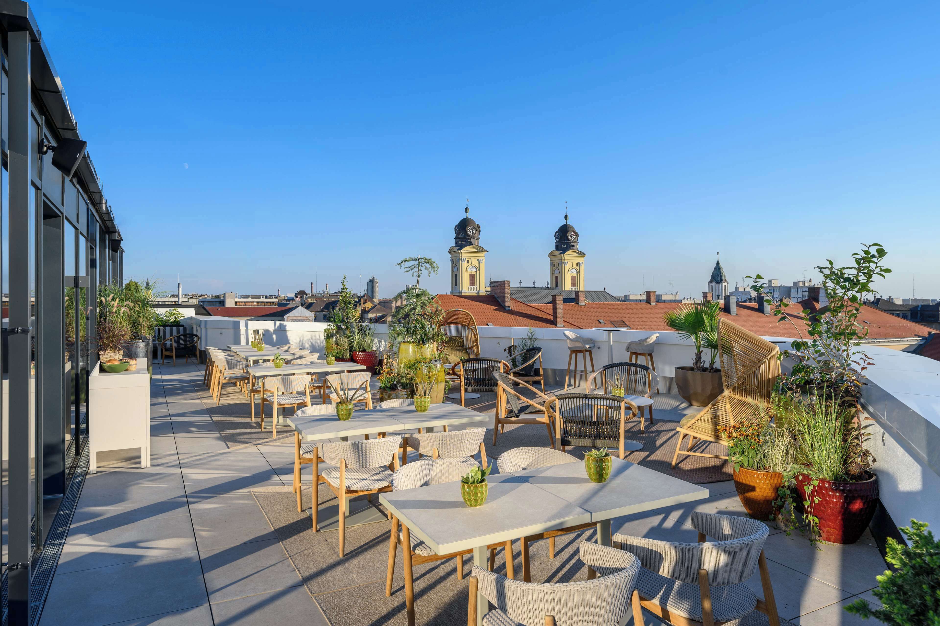 A rooftop terrace with outdoor seating, potted plants, and a view of city buildings and towers.