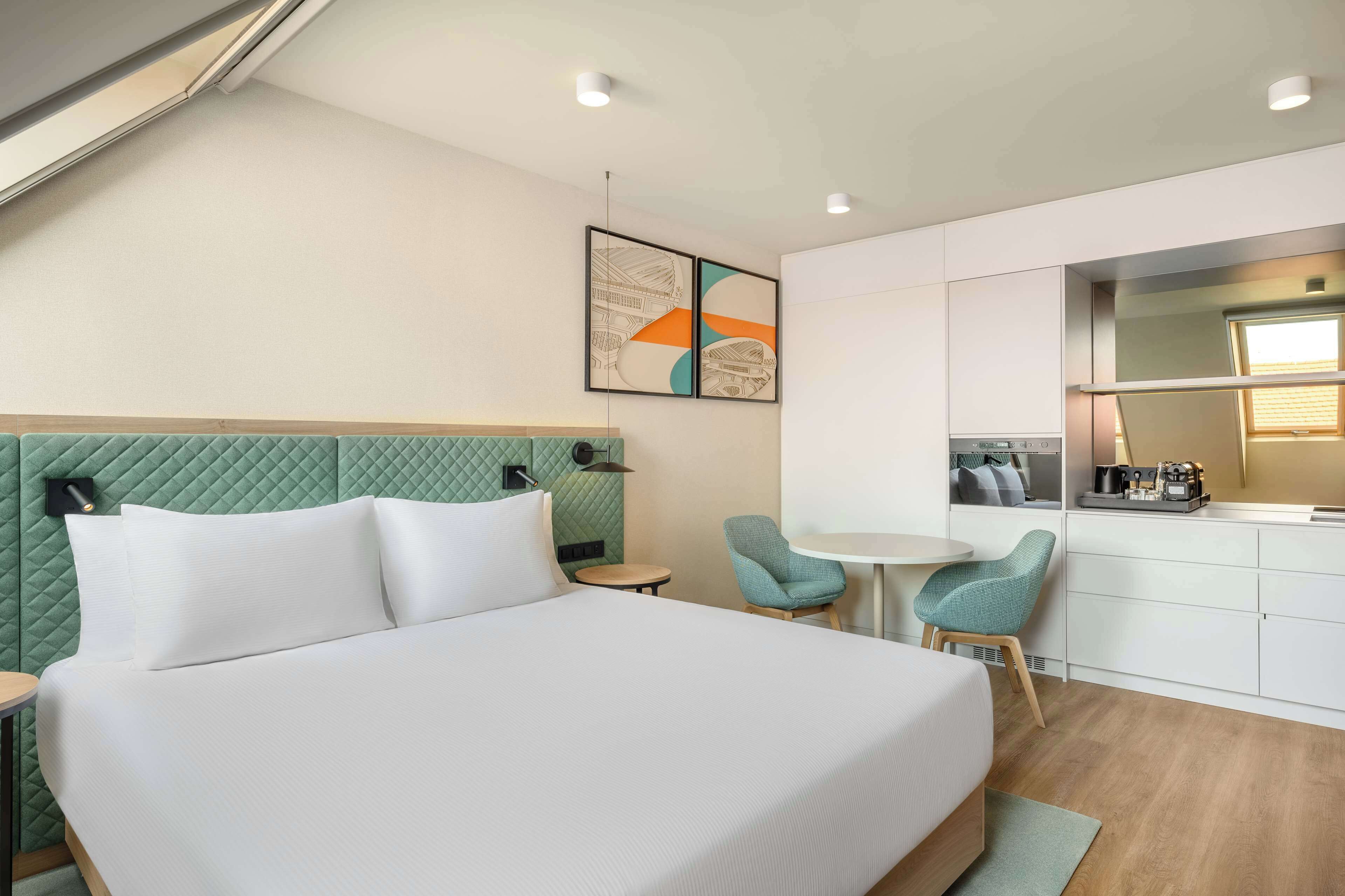 A modern hotel room with a bed, a small dining table, two chairs, a kitchenette, and a green accent wall.