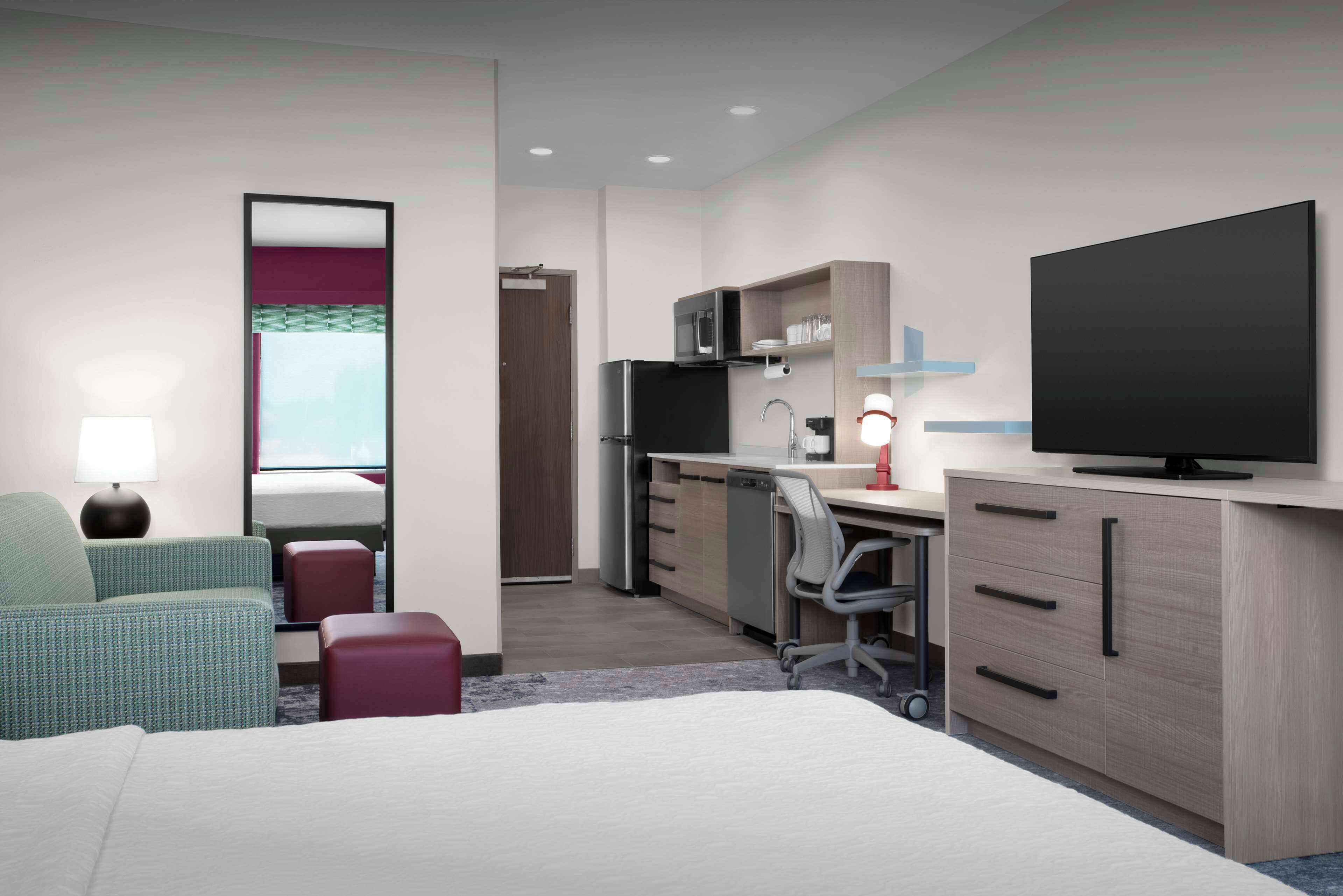 A modern hotel room with a bed, a desk with a computer, a flat-screen TV, a refrigerator, and a microwave.