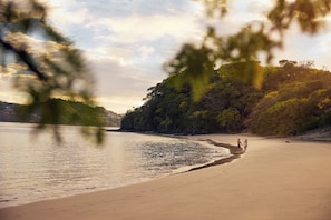 A beach with a couple walking, trees, and a calm sea. - Waldorf Astoria Costa Rica Punta Cacique (Sardinal)