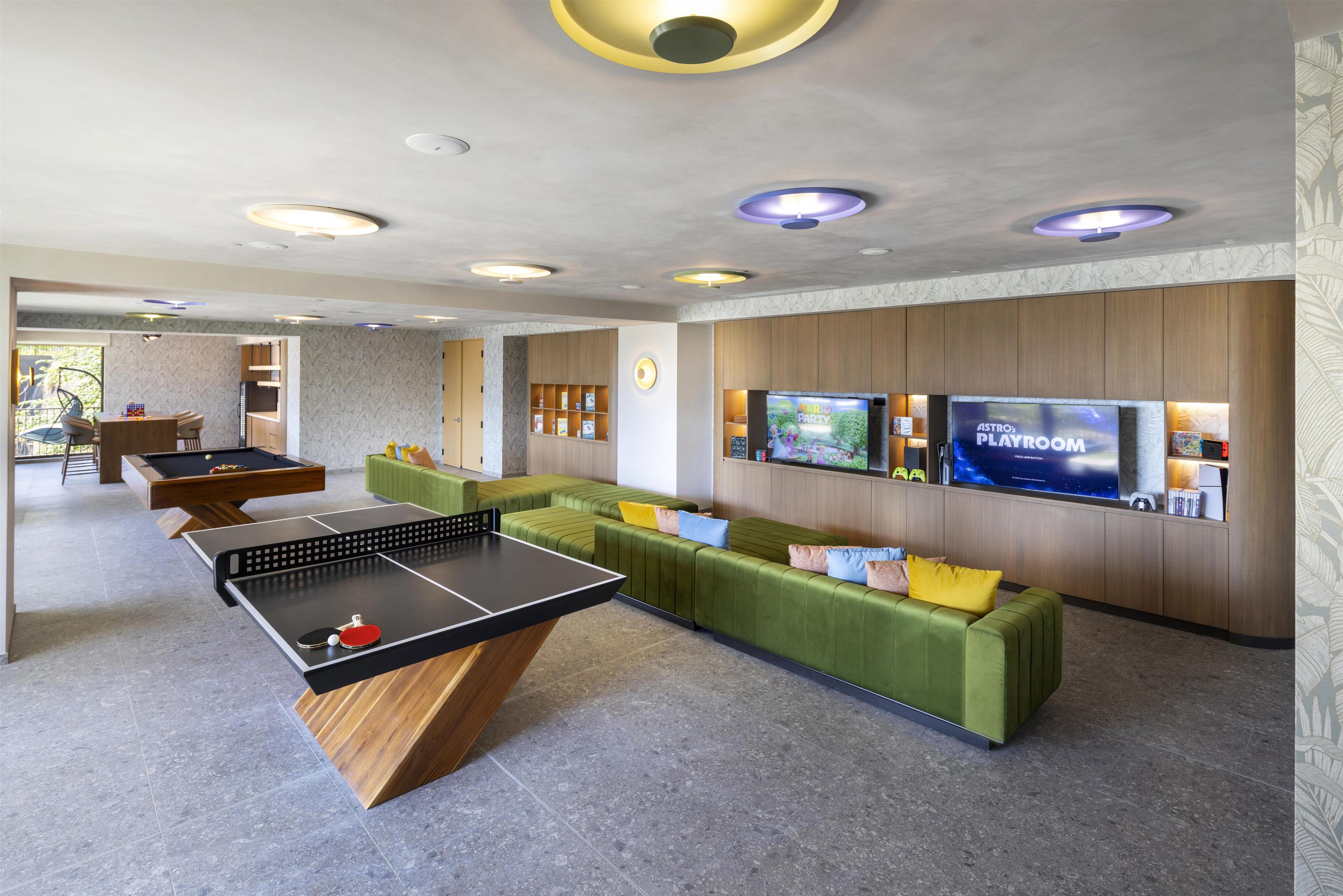 A modern lounge area with a ping pong table, green sofas, and a flat-screen TV displaying 'ASTRO'S PLAYROOM'.