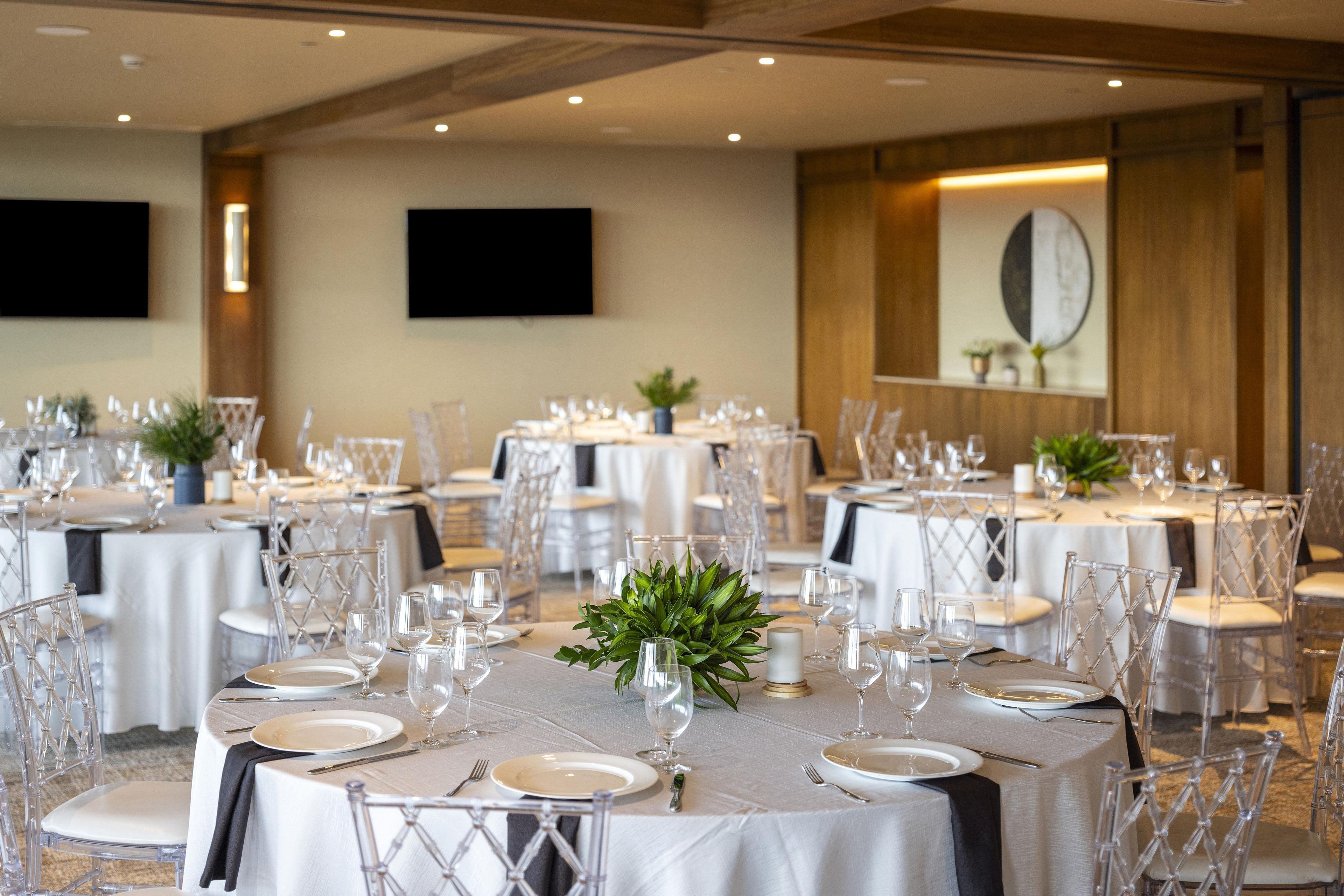 Un salón de banquetes con mesas redondas dispuestas para comer, sillas transparentes y un televisor montado en la pared.