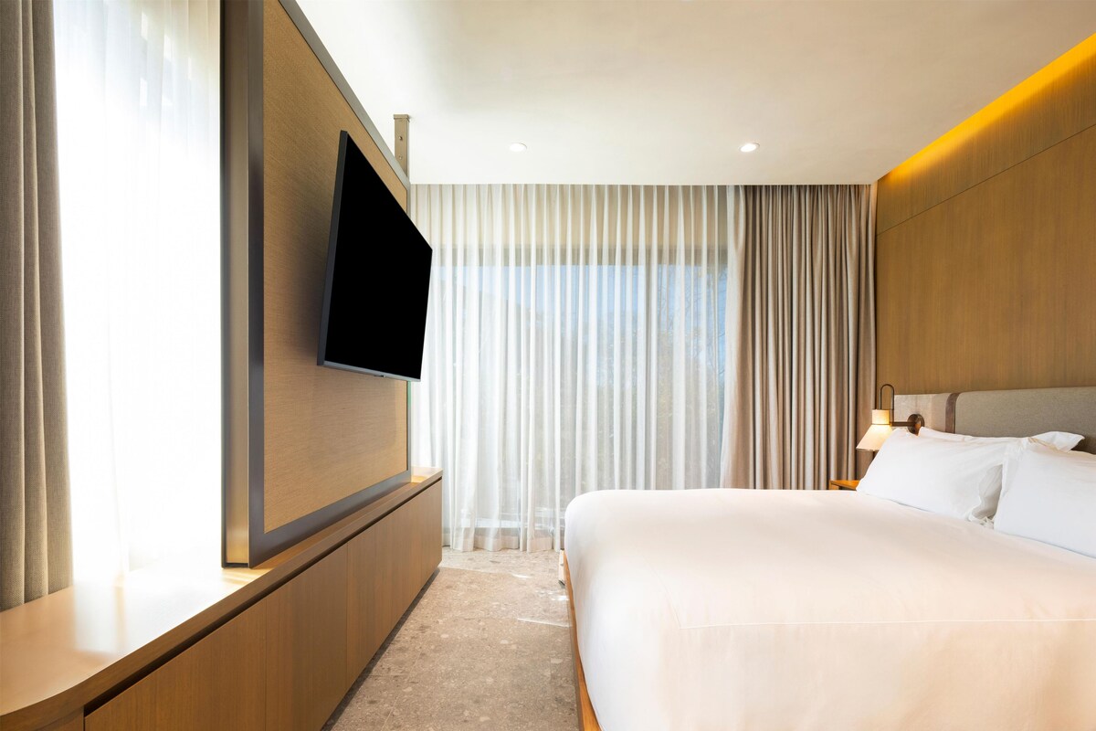 A hotel room with a bed, a TV mounted on the wall, and a large window with curtains.