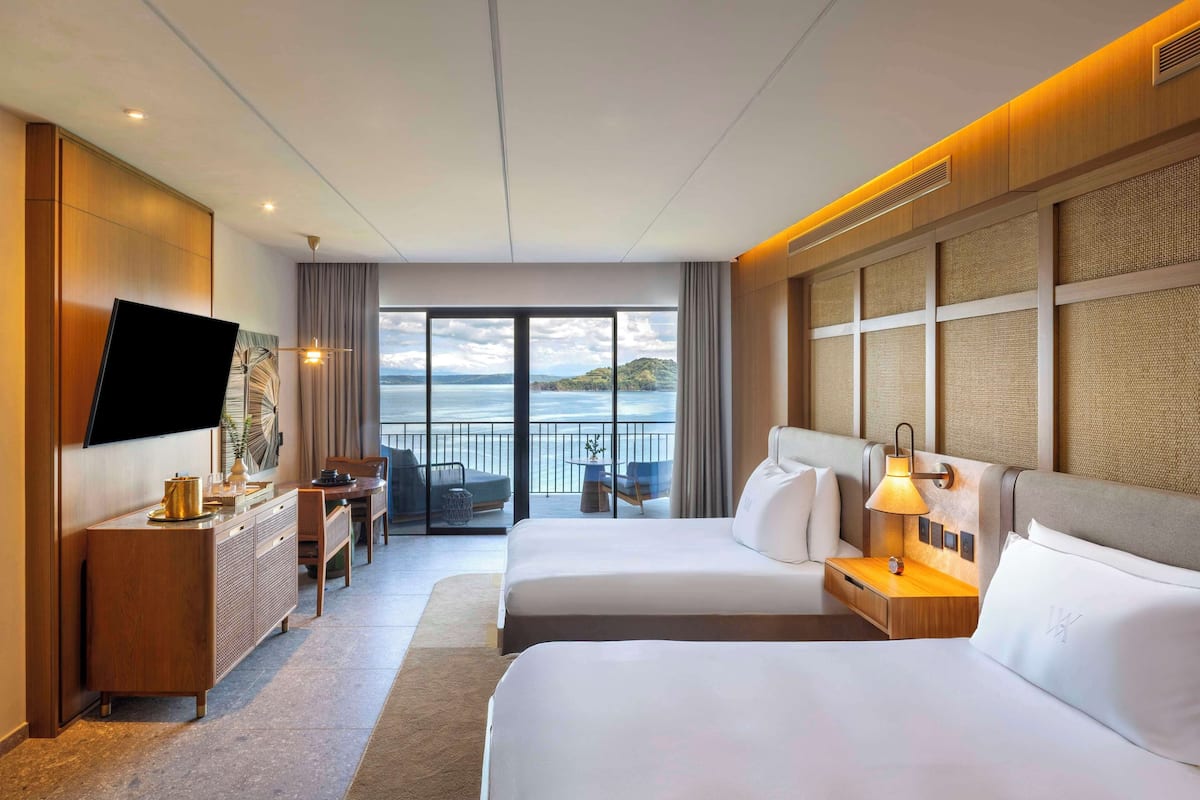A hotel room with two beds, a TV, a desk, and a view of the sea.