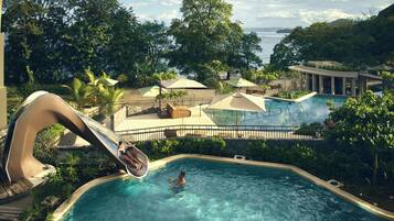 A pool area with a slide, lounge chairs, umbrellas, and a view of the sea.