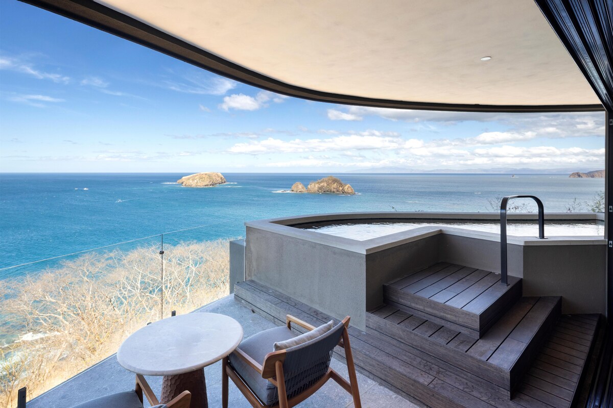 A balcony with a view of the ocean, a hot tub, and a table with two chairs.