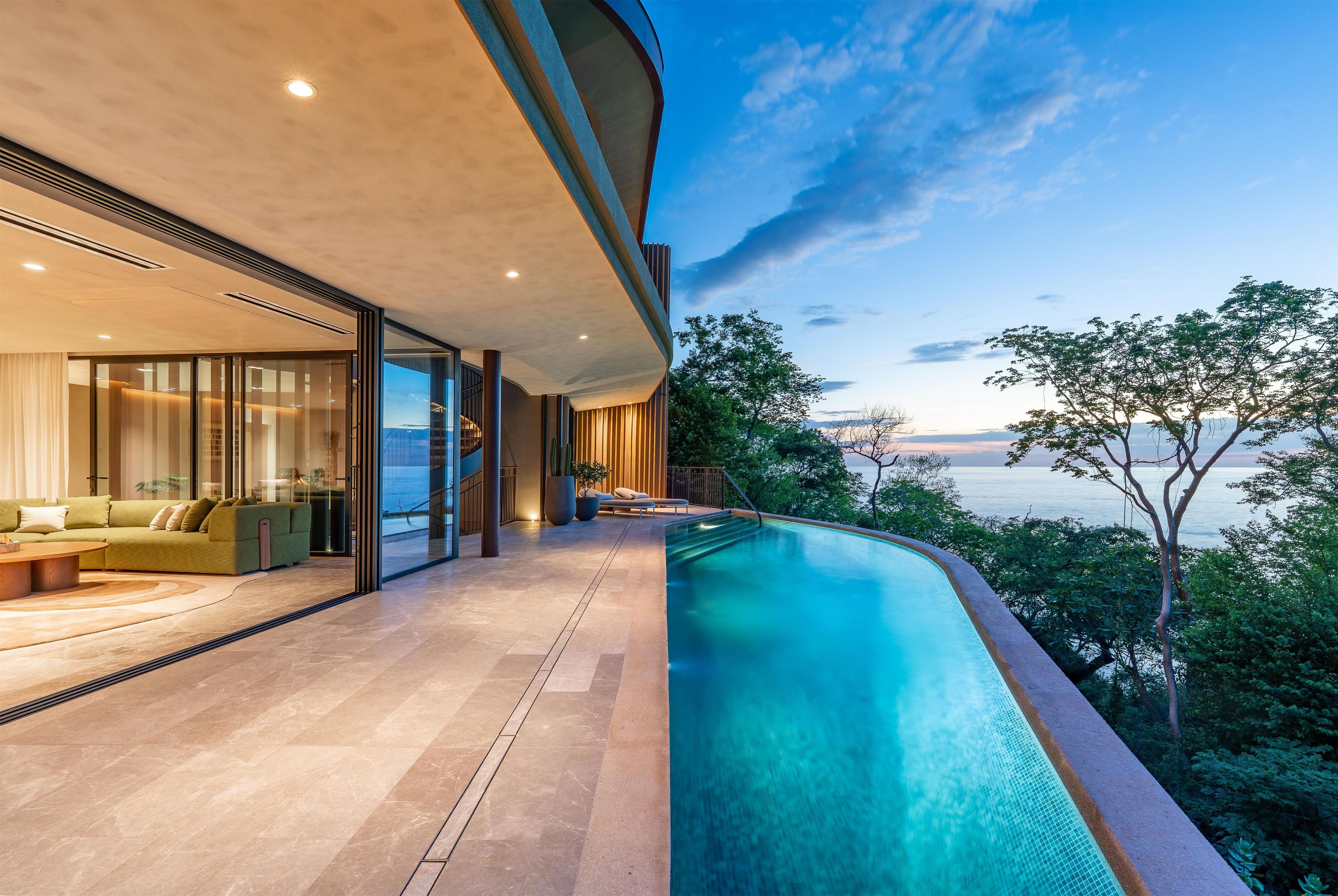 Ein moderner Poolbereich im Freien mit Meerblick, eine Lounge mit Sofa und Couchtisch sowie ein gläserner Raum mit Blick ins Innere.