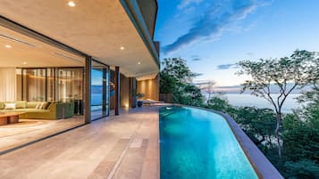 Ein moderner Poolbereich im Freien mit Meerblick, eine Lounge mit Sofa und Couchtisch sowie ein gläserner Raum mit Blick ins Innere.