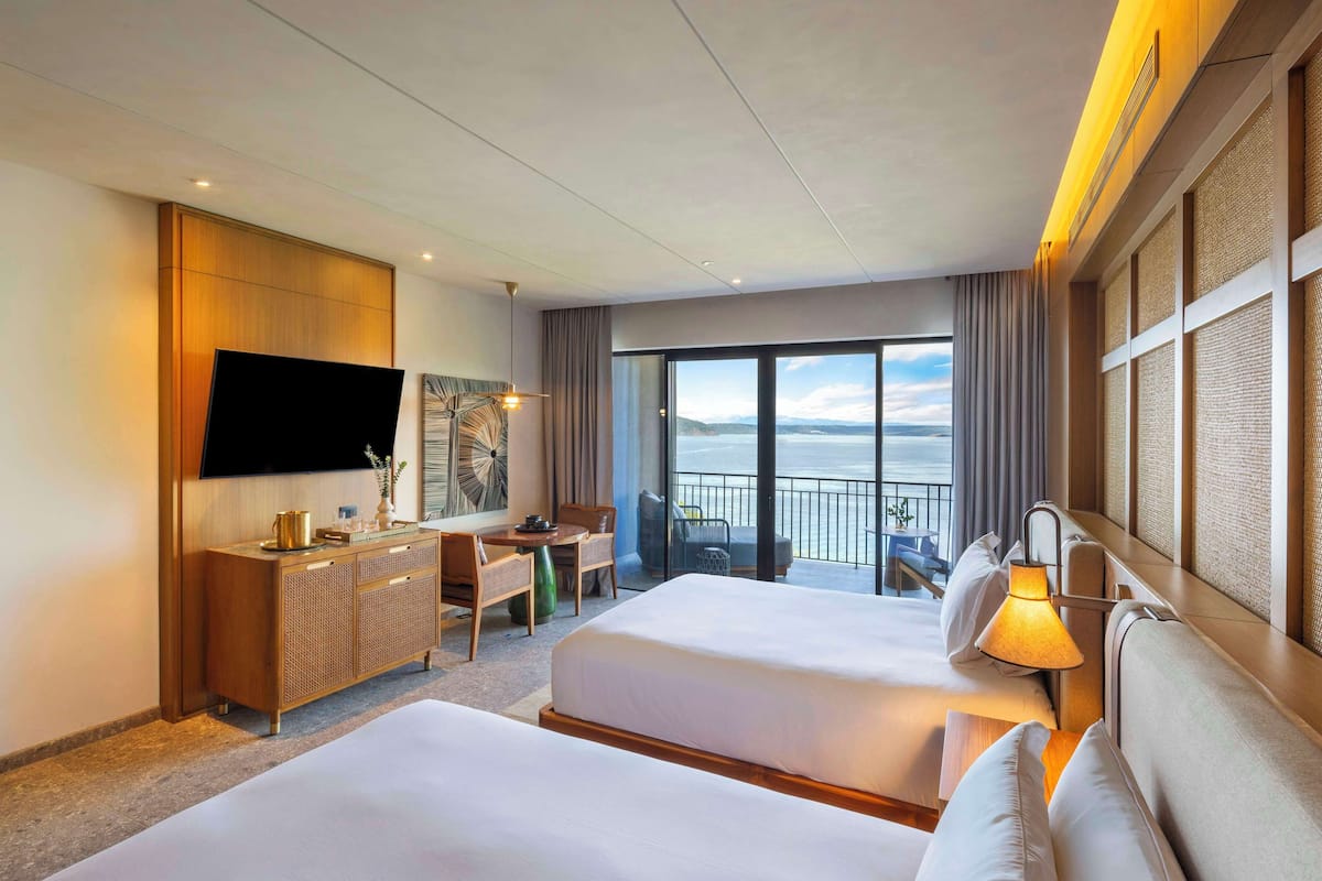 A hotel room with two beds, a desk, a television, and a balcony with a view of the sea.