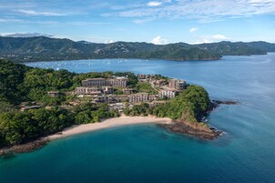 Vista aérea de un complejo turístico costero con varias edificaciones, una playa de arena y una bahía con barcos.