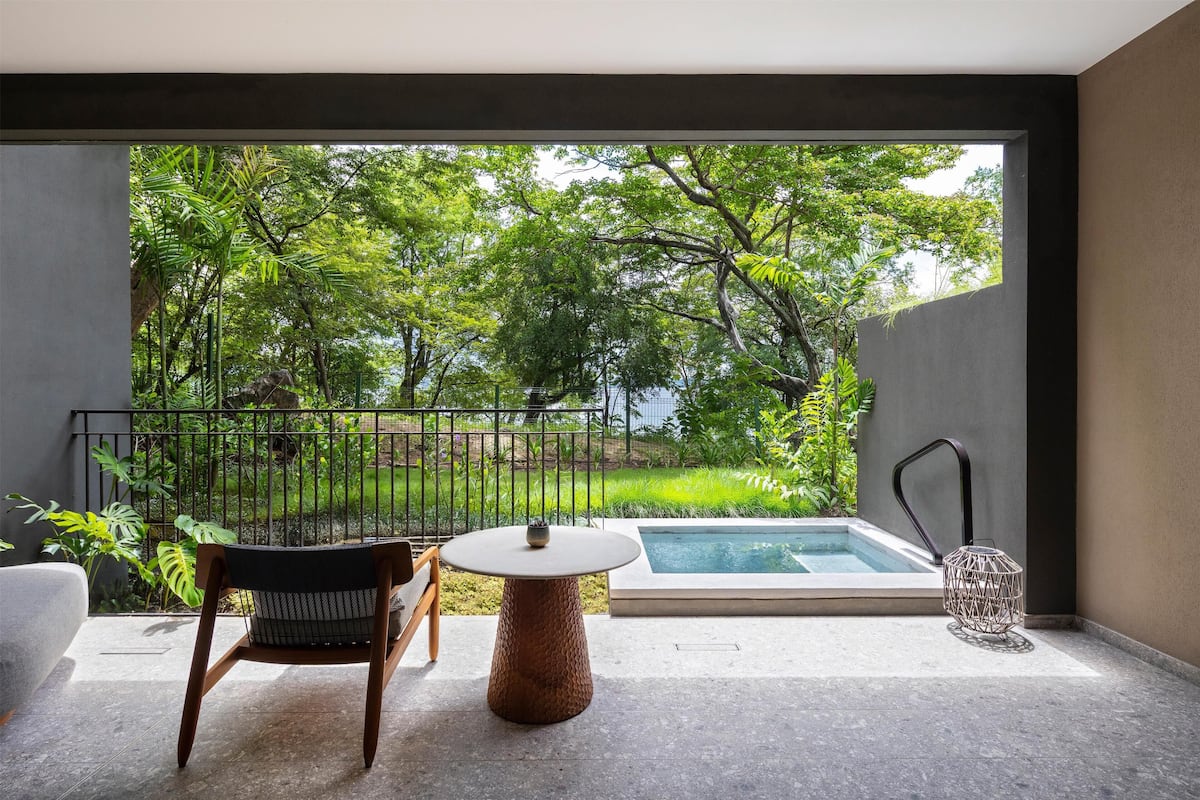 A modern outdoor patio with a round table and chairs, a small pool, and lush greenery visible through a large window.