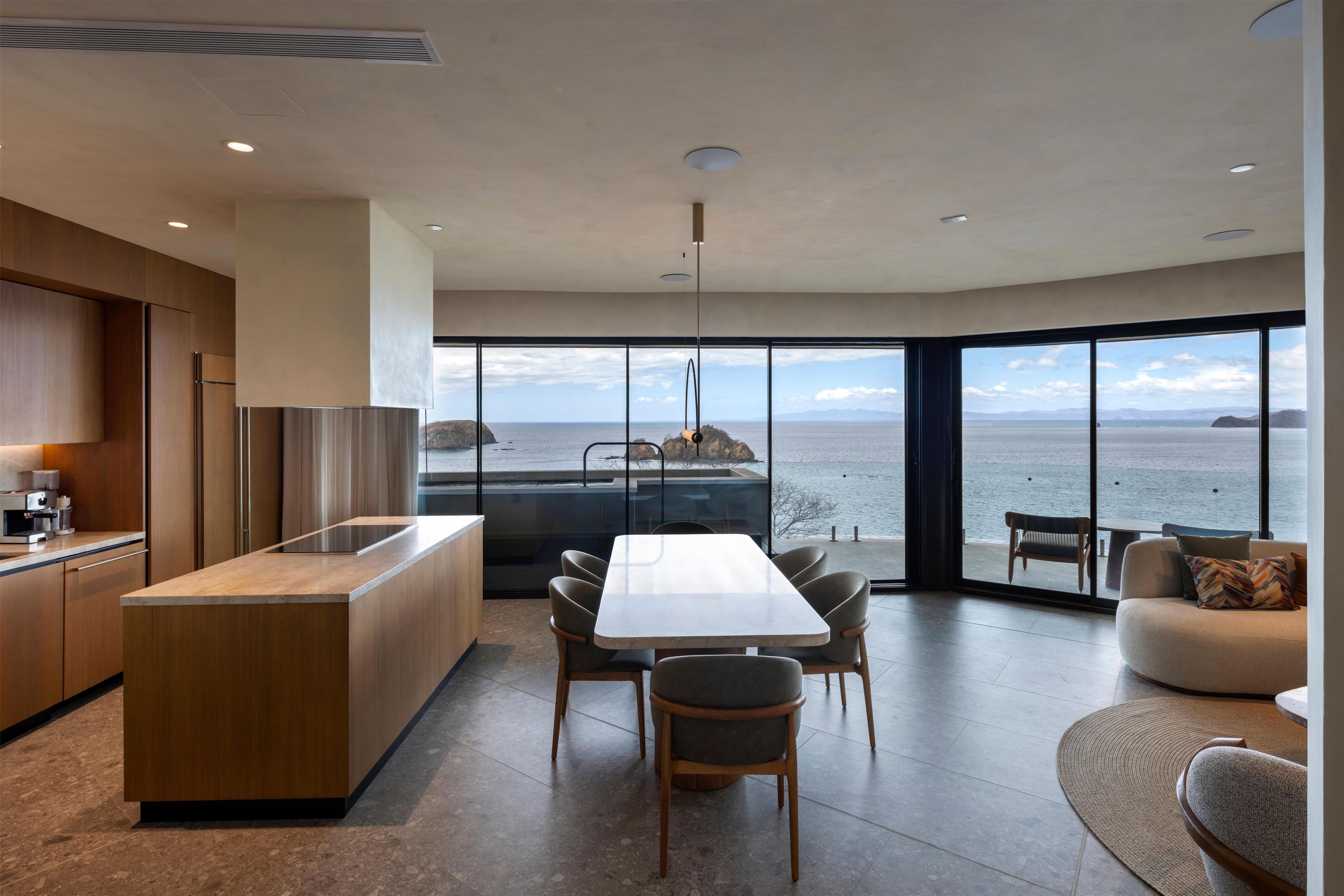 Una cocina moderna con una isla grande, comedor con sillas y vista al mar a través de amplios ventanales.