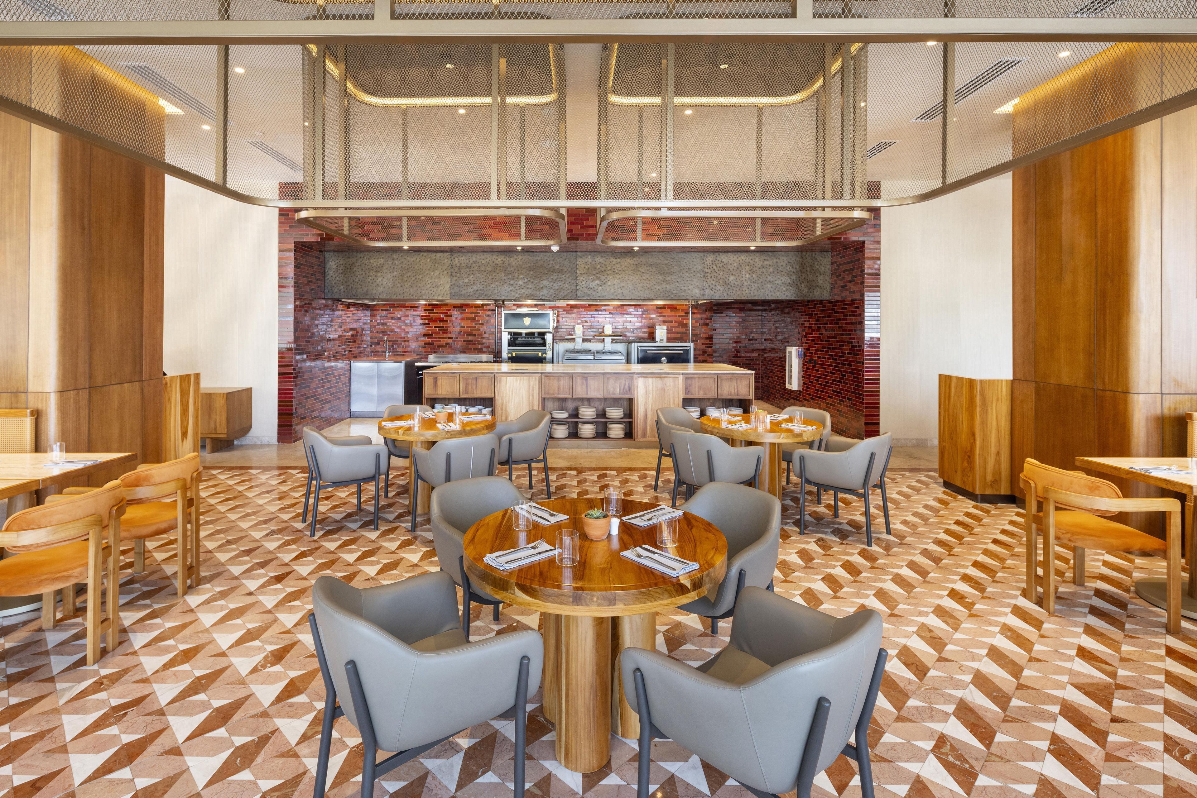 Un comedor moderno con mesas y sillas redondas, una pared de ladrillo con una parrilla y un techo con un diseño geométrico.