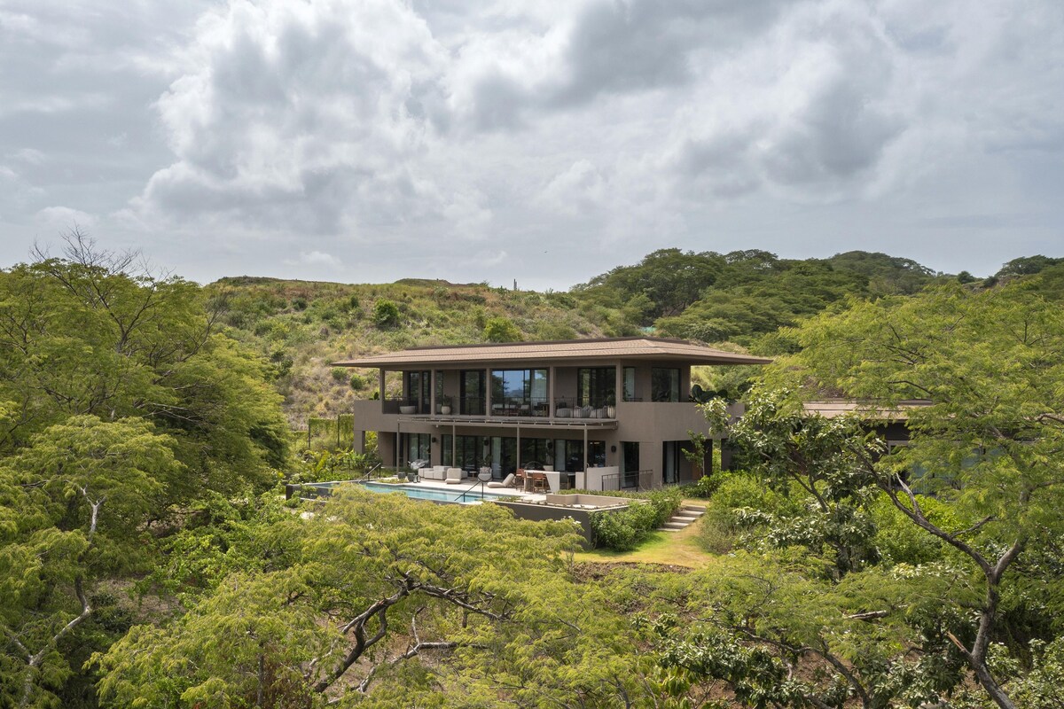 A modern two-story house with a swimming pool, surrounded by lush greenery and hills.