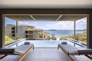 A modern hotel room with a glass infinity pool, two lounge chairs, and a view of the sea and buildings. - Waldorf Astoria Costa Rica Punta Cacique (Sardinal)
