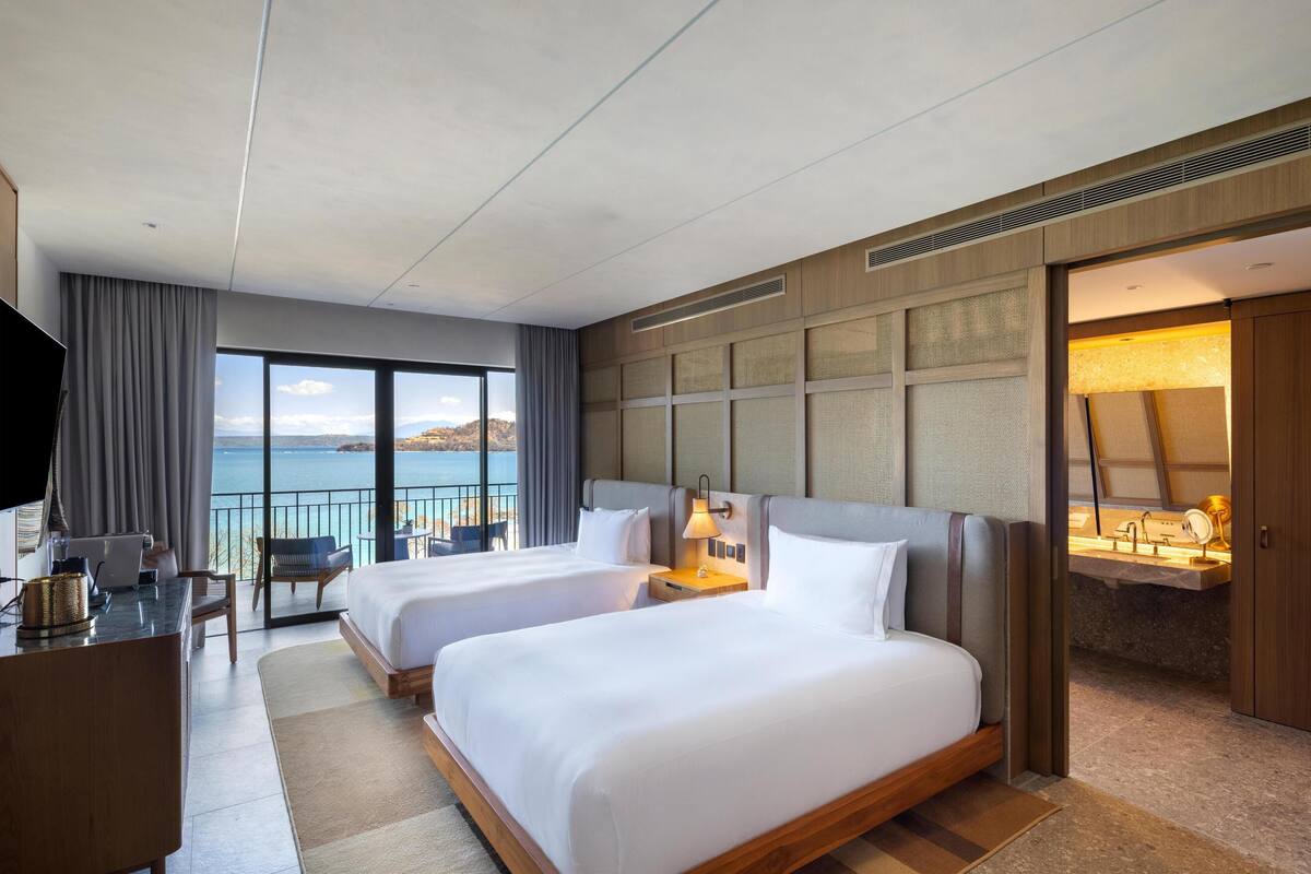 A hotel room with two beds, a desk, a chair, a TV, and a view of the sea.