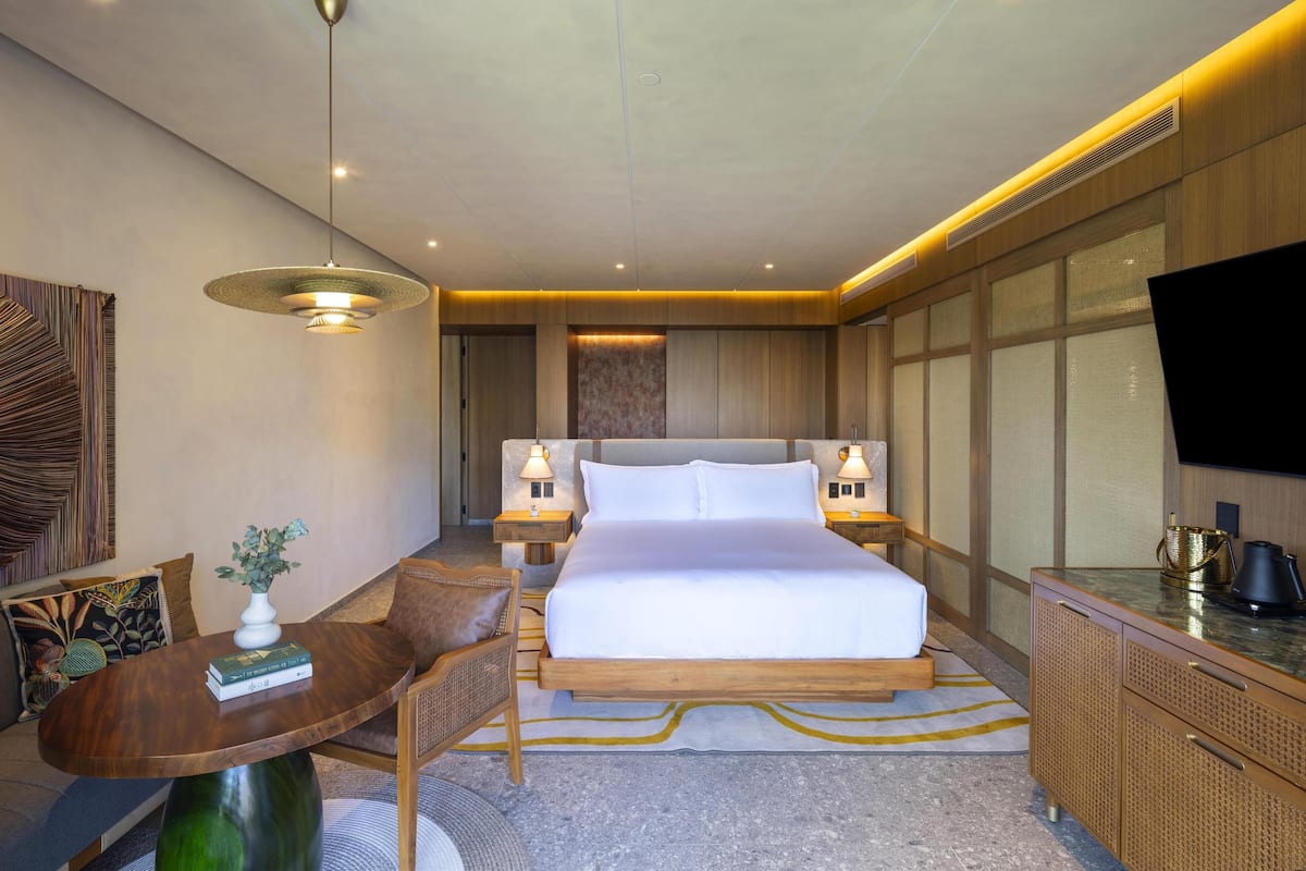 A modern hotel room with a large bed, wooden headboard, bedside lamps, a flat-screen TV, and a dining area with a table and chairs.