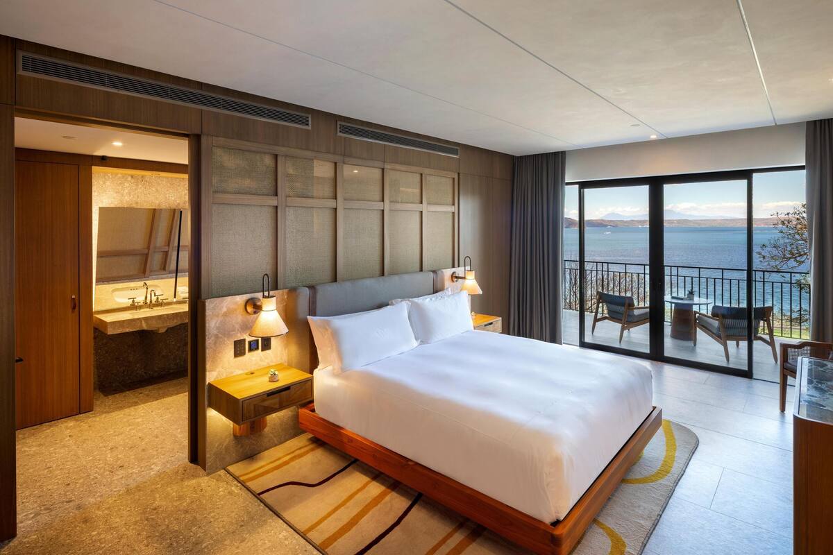 A modern hotel room with a large bed, a bathroom with a vanity, and a balcony with a view of the sea.