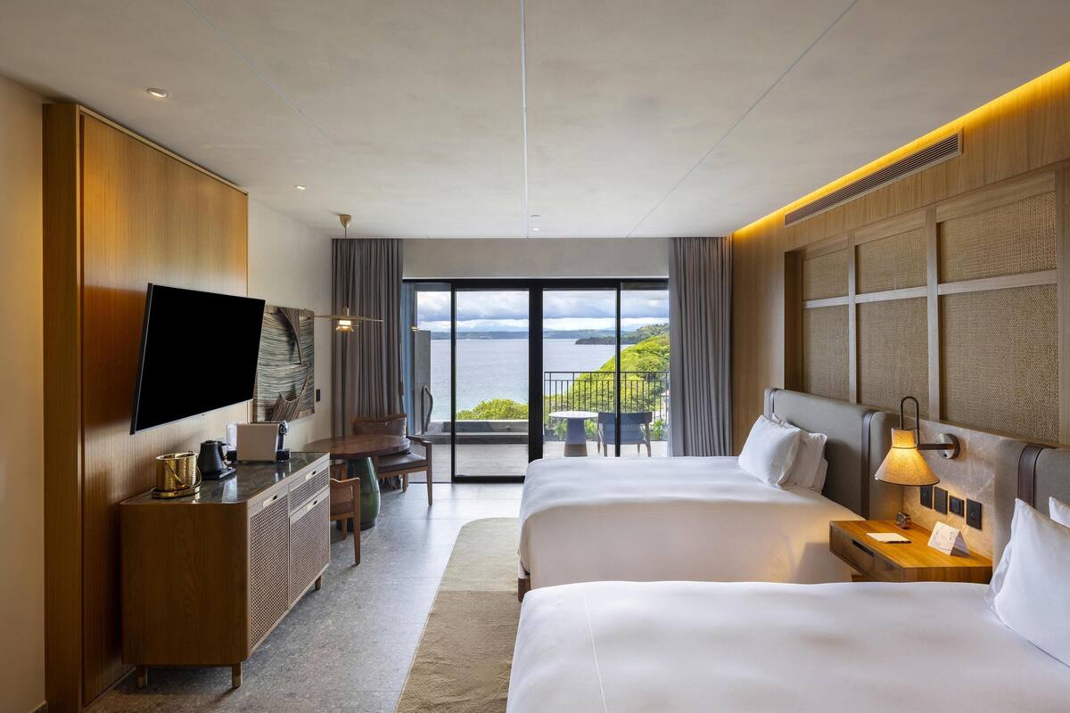 A hotel room with two beds, a TV, a desk, and a balcony with a view of the sea.