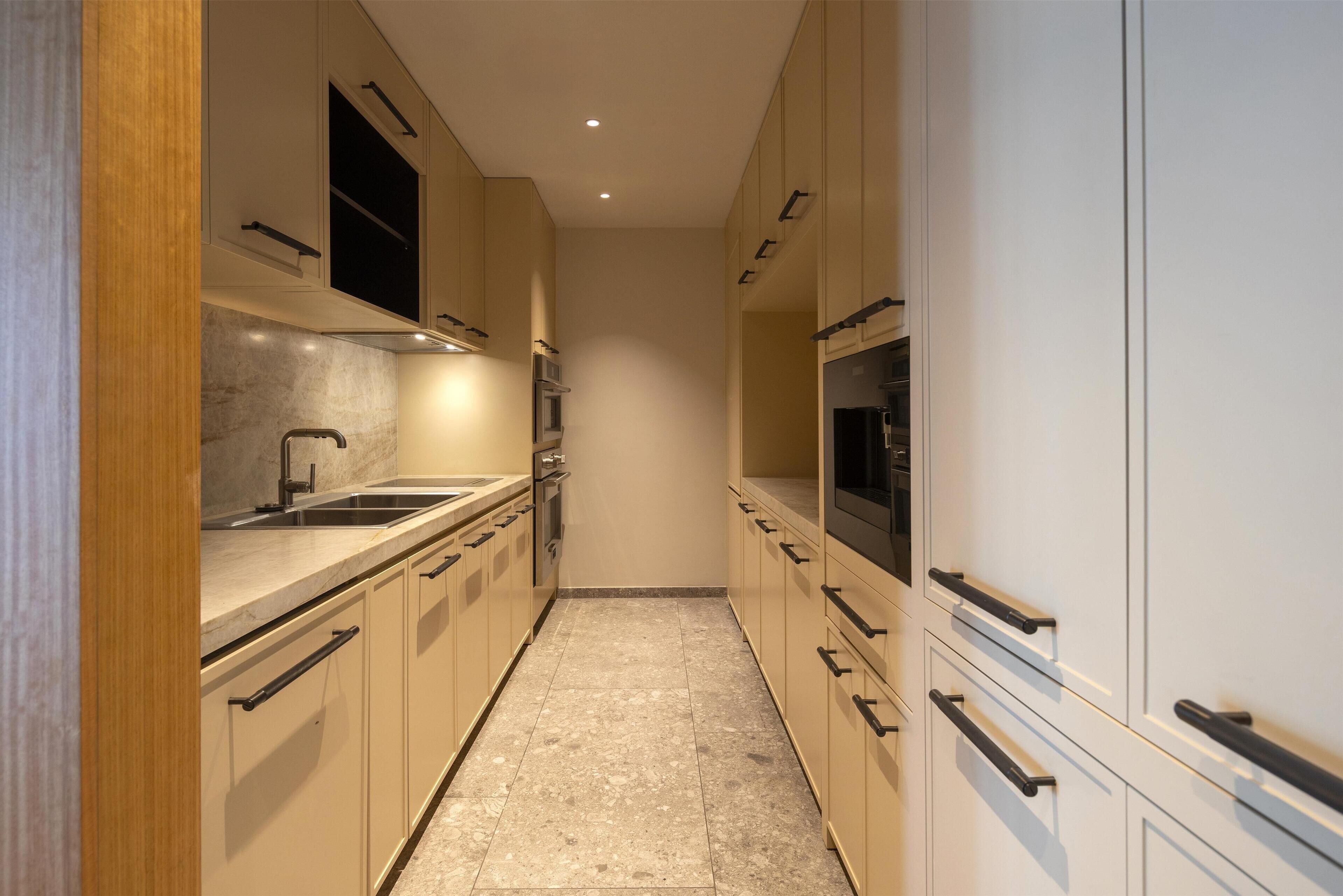 A narrow kitchen with beige cabinets, a marble countertop, and a black oven.