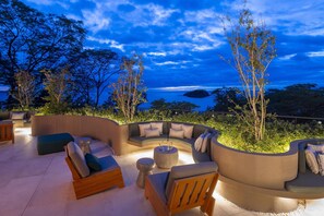 An outdoor seating area with a curved sofa, round ottoman, and wooden chairs, overlooking a scenic view of the ocean and trees. - Waldorf Astoria Costa Rica Punta Cacique (Sardinal)