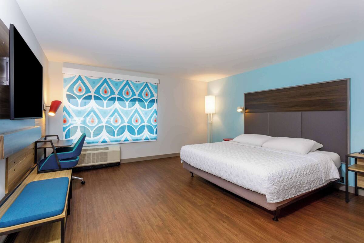 A hotel room with a bed, a desk with a chair, a TV, and a window with a patterned curtain.