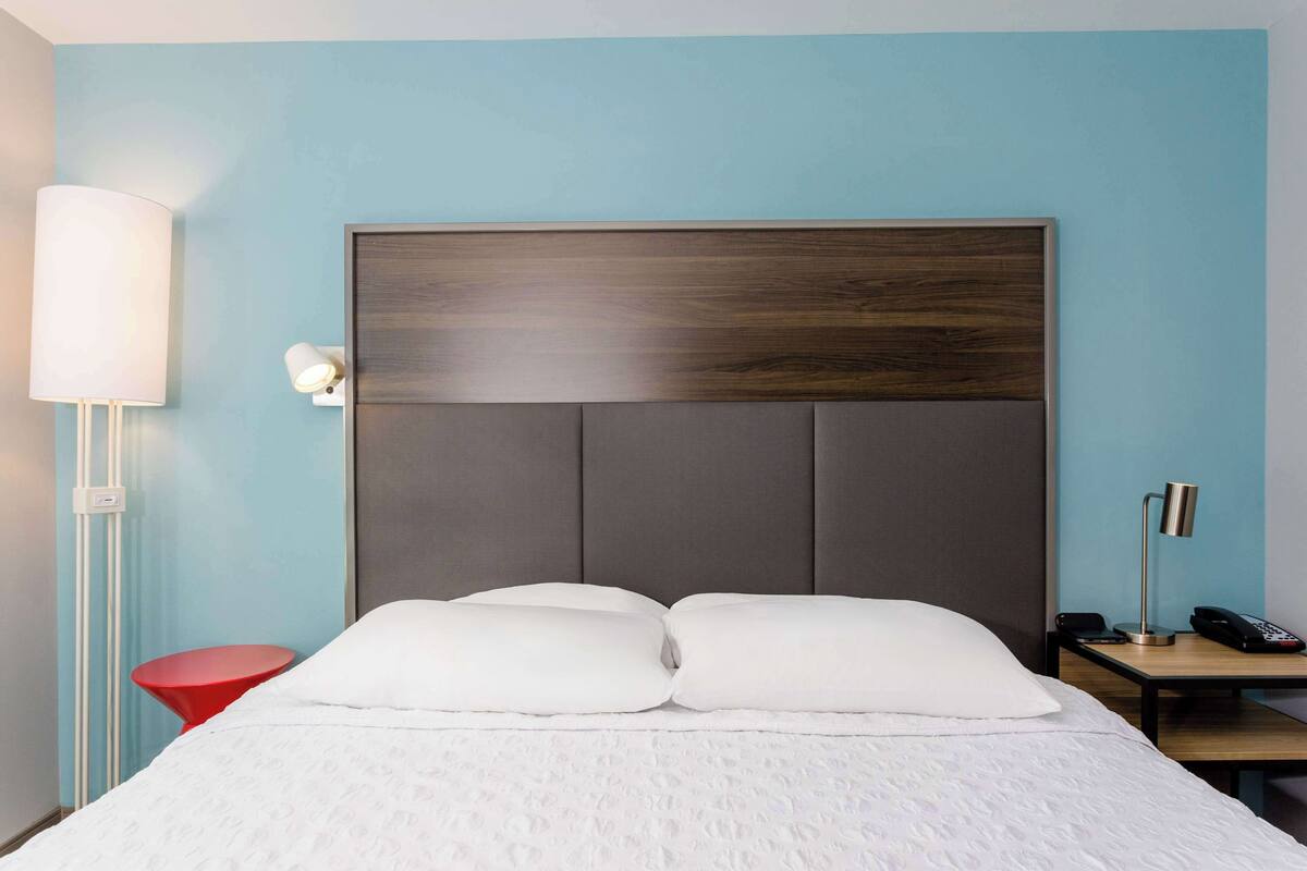 A hotel room with a large bed, a bedside table with a lamp, a red chair, and a nightstand with a phone.