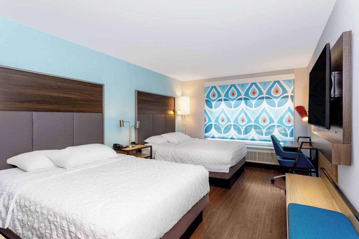 A hotel room with two beds, a desk, a chair, a TV, and a patterned wall.