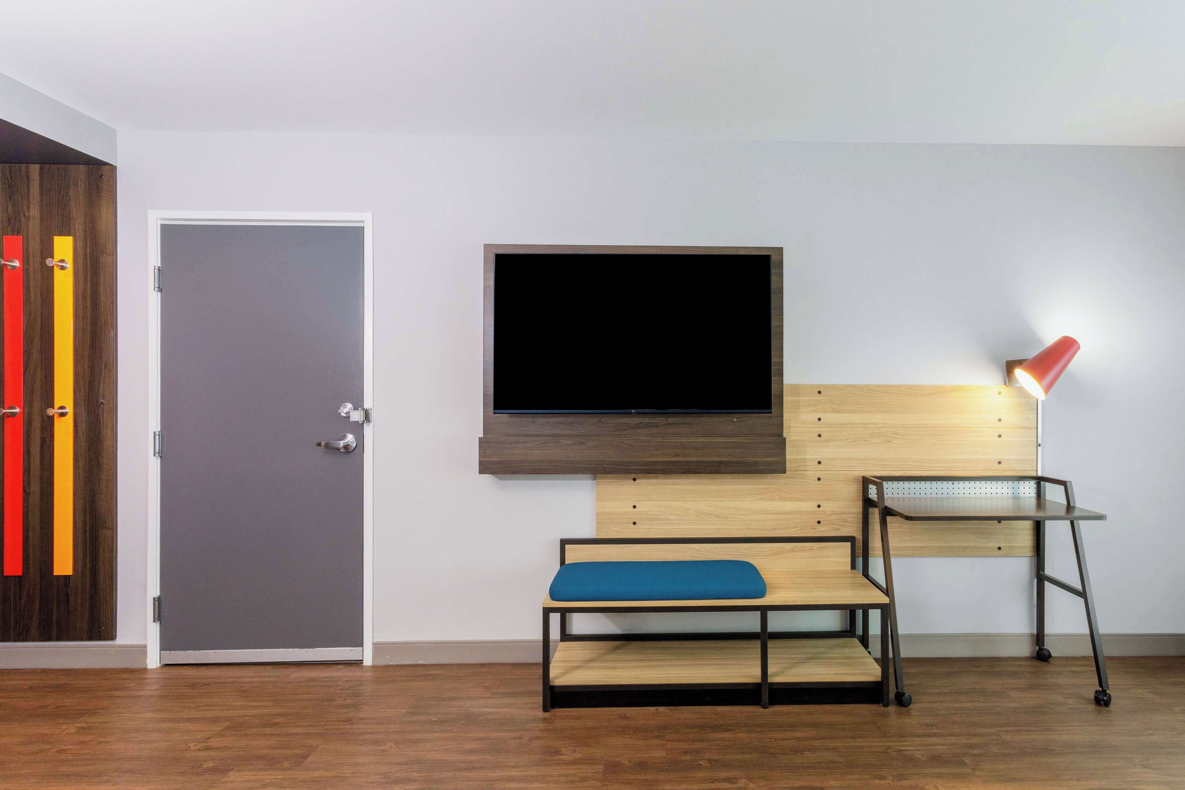 A room with a wooden floor, a grey door, a wall-mounted TV, a bench with a blue cushion, and a desk with a red lamp.