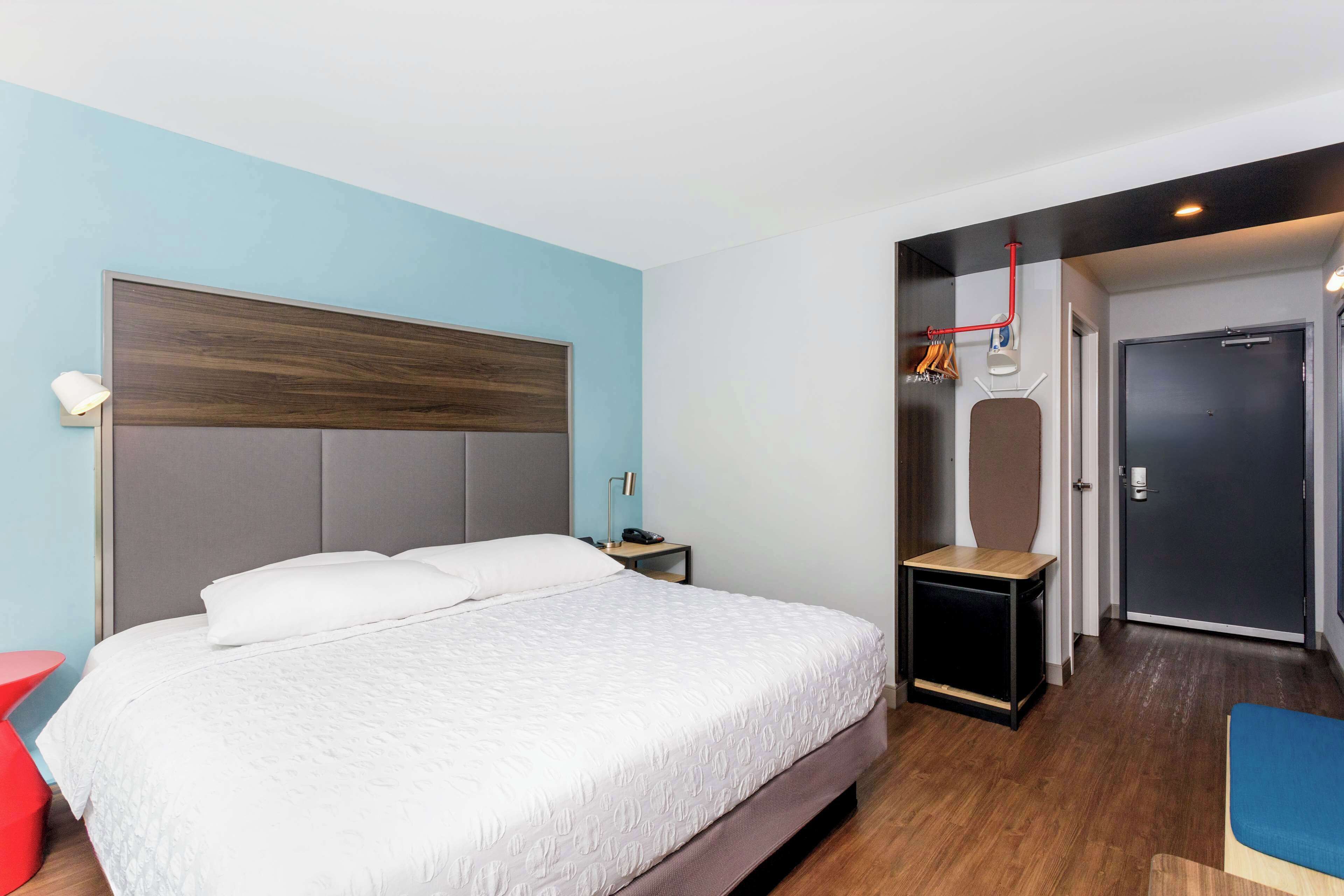 Une chambre d’hôtel moderne avec un grand lit, une table de chevet, une chaise rouge et une armoire avec un miroir.