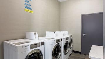 Un lavadero con tres lavadoras y secadoras industriales blancas, una puerta gris e indicaciones para lavar en la pared.