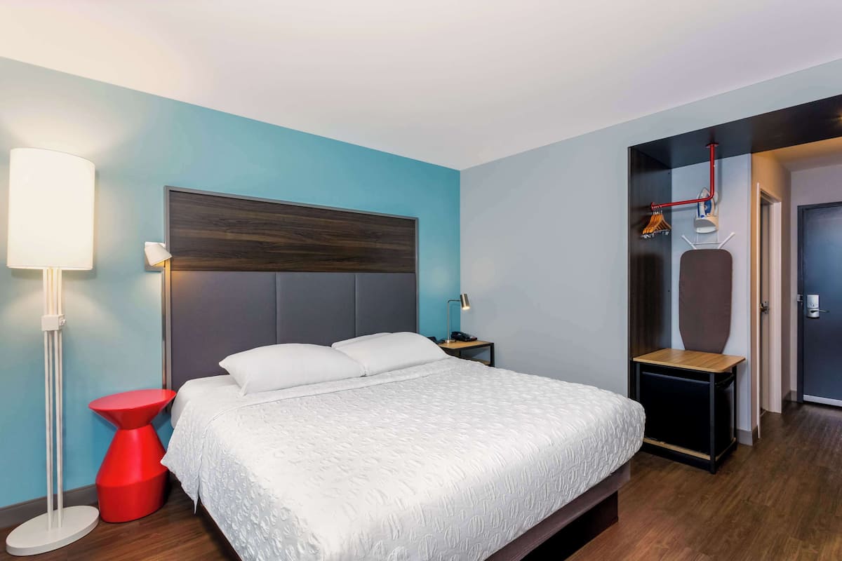 A modern hotel room with a large bed, a red side table, a floor lamp, and a desk with a chair.