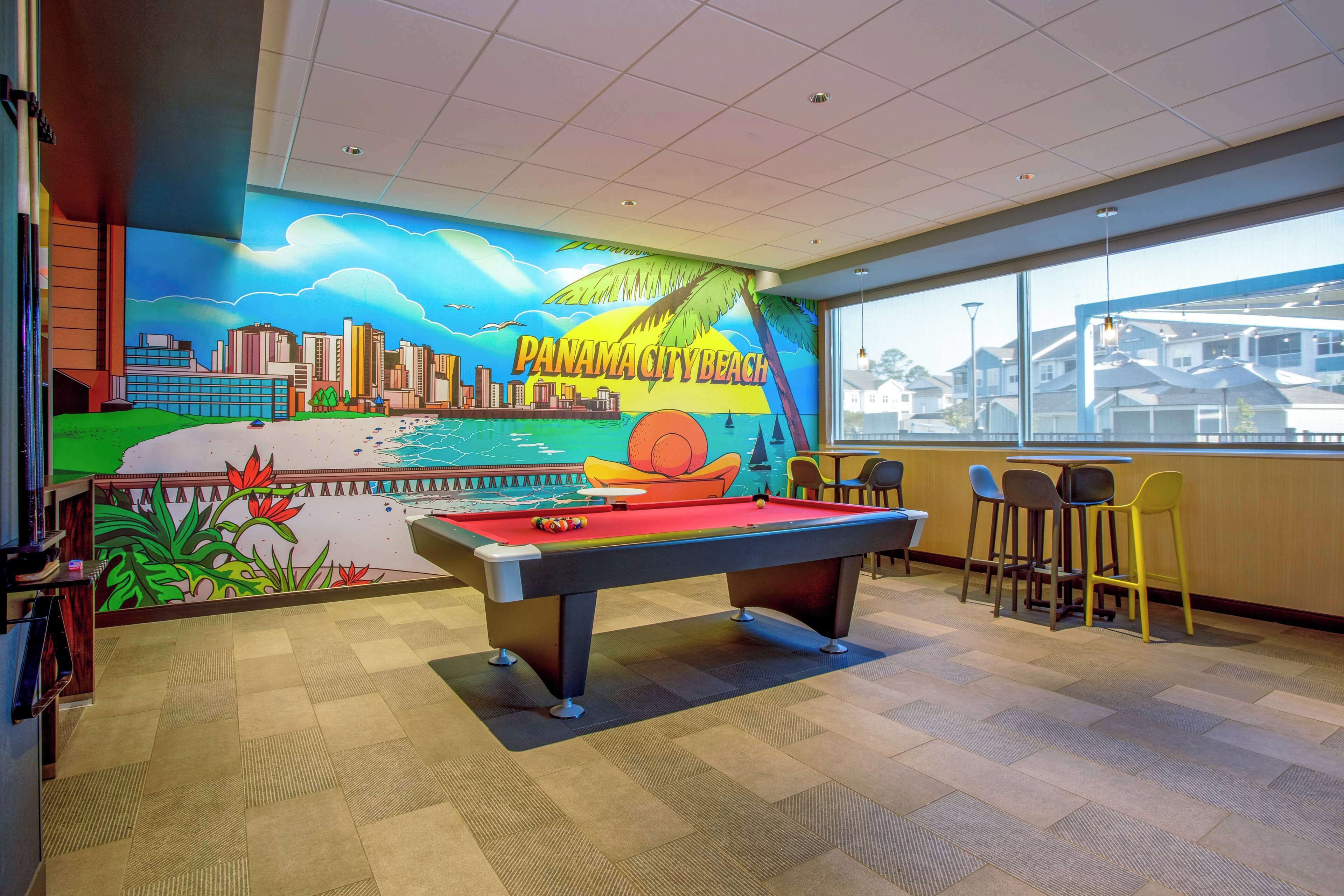 A pool table with a mural of Panama City Beach, a bar area with chairs, and a view of houses through large windows.